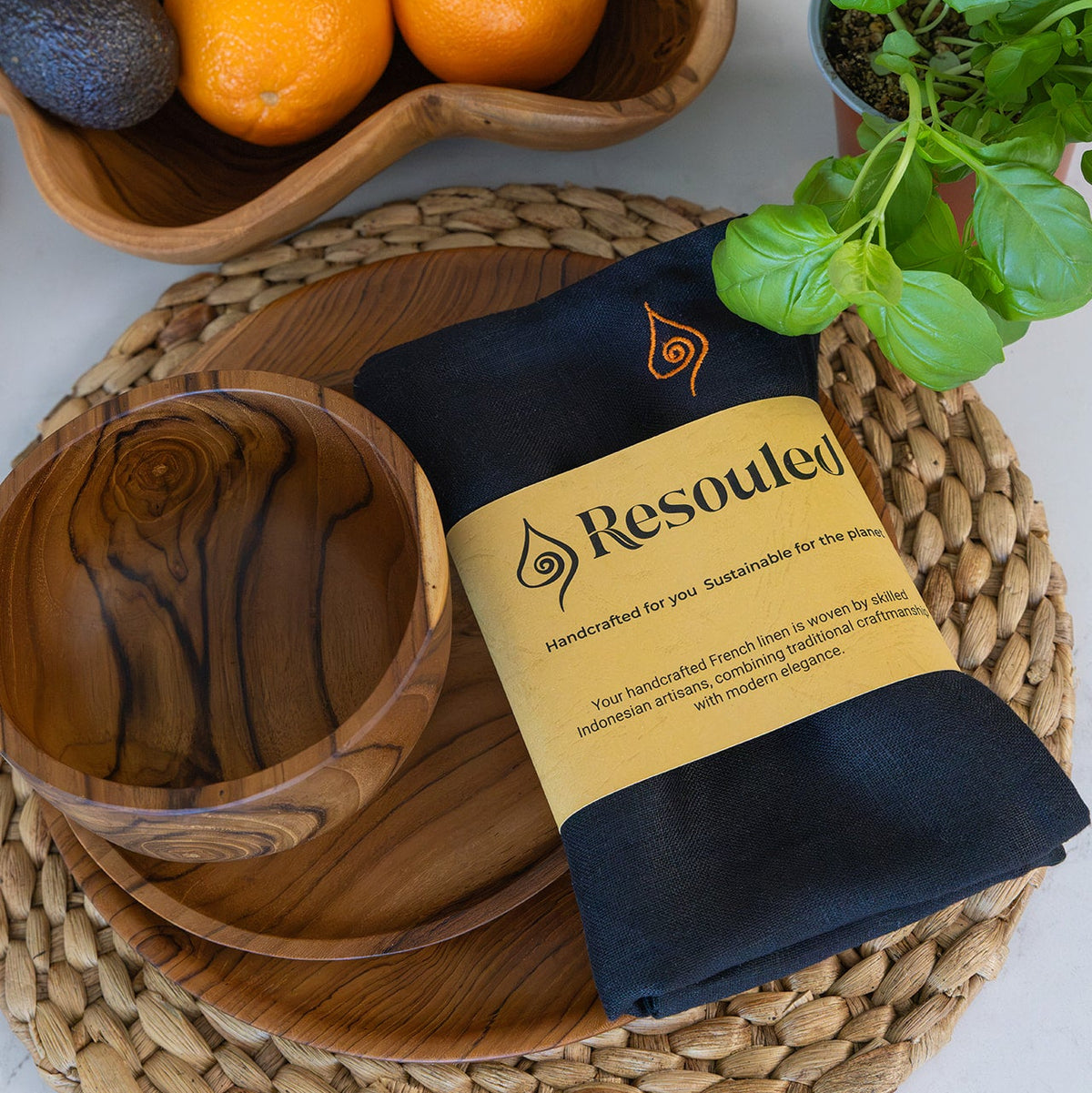 A folded black French Linen cloth napkin with a tan paper label reading 'Resouled' is arranged on teak wooden plates on a woven placemat beside a wooden bowl and a potted basil plant. A fruit bowl appears in the background.