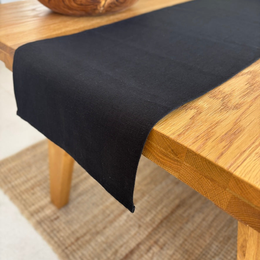 Image shows folded black table runner on a set dining table. Hand-woven from 100% sustainable French linen, this table runner adds an earthy elegance and style to your dining table. Dimensions: 220cm x 30cm