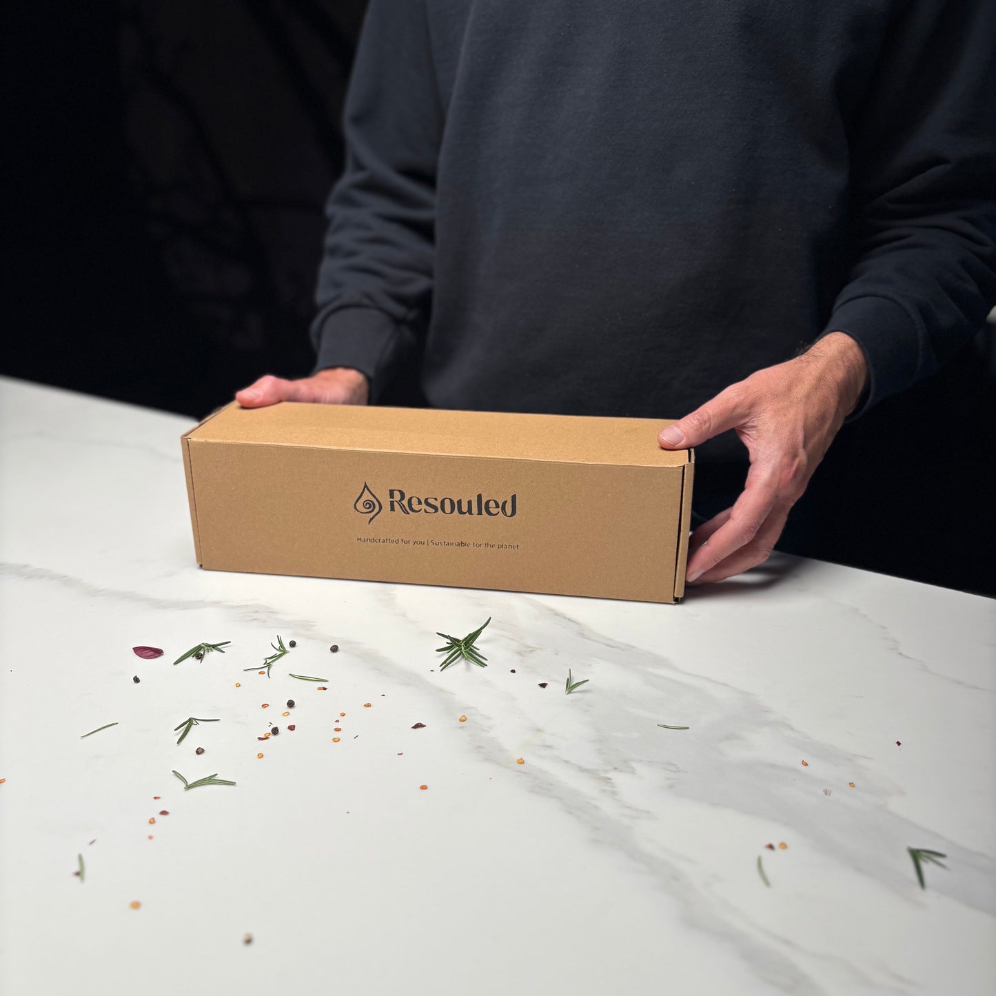 Person holding a cardboard box with 'Resouted' branding on a white surface with scattered herbs.