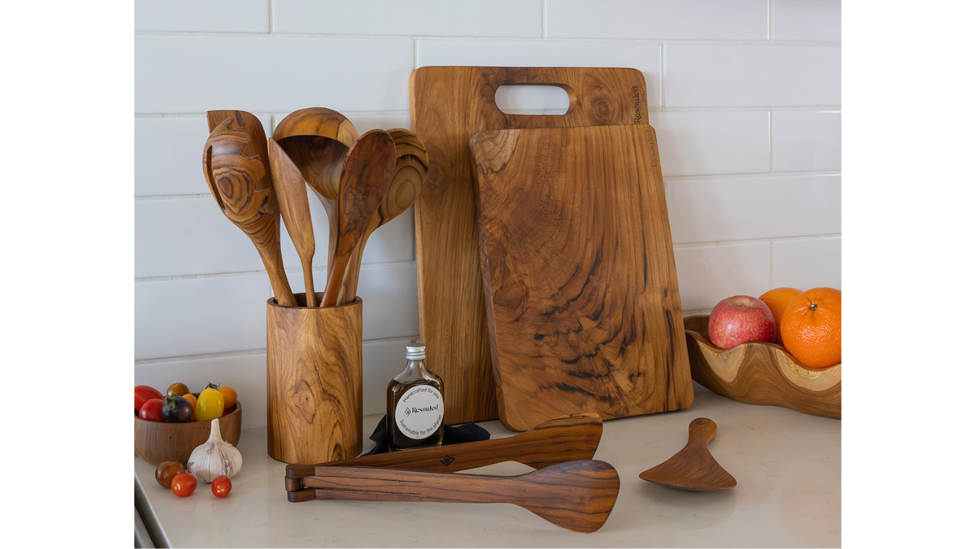 A set of handcrafted teak wood kitchen utensils, including teak chopping boards, rice spoon, and Utensil set, arranged on a light countertop against a white subway tile wall.