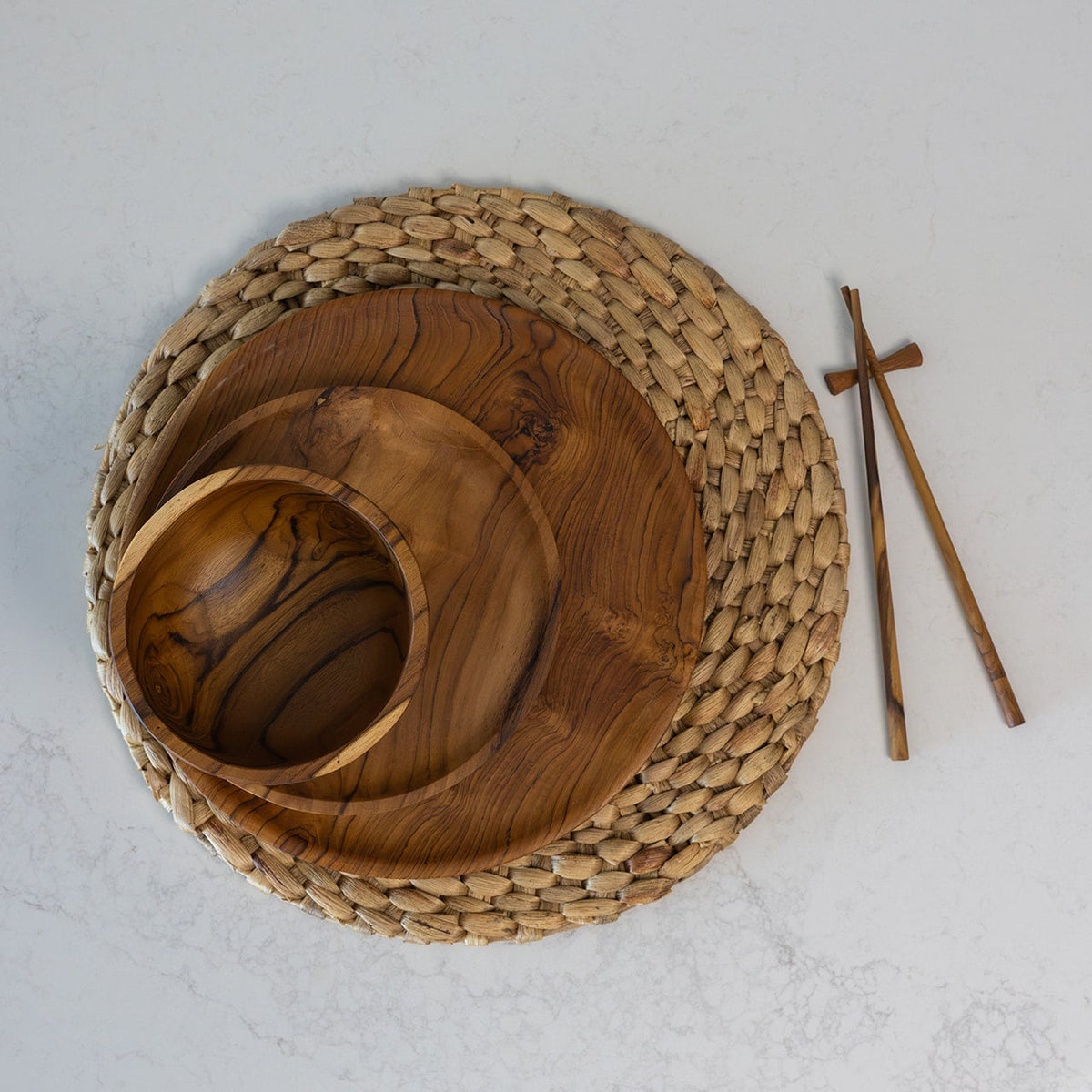 A teak wooden bowl, on top of a small wooden plate, on top of a larger wooden plate, all with a prominent wavy grain pattern, arranged on a round woven seagrass placemat with wooden chopsticks on a light gray marble surface.