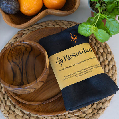 A folded black French Linen cloth napkin with a tan paper label reading 'Resouled' is arranged on teak wooden plates on a woven placemat beside a wooden bowl and a potted basil plant. A fruit bowl appears in the background.