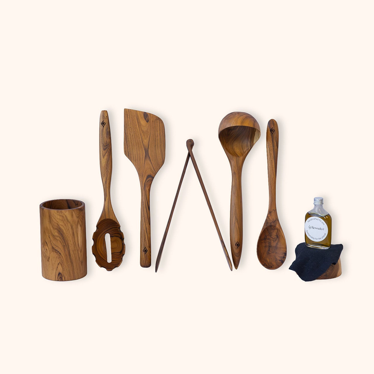 A 6 piece set of sustainable, handcrafted teak kitchen utensils including wooden spoon, spatula, spaghetti spoon, ladle and tongs, displayed next to a small bottle of food safe hemp oil on a light cream background.