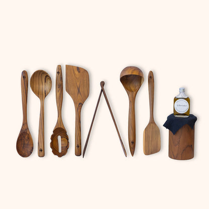 An 8 piece set of sustainable, handcrafted teak kitchen utensils including wooden spoon, spatula, spaghetti spoon, ladle, serving spoon, turner and tongs, displayed next to a small bottle of food safe hemp oil on a light cream background.