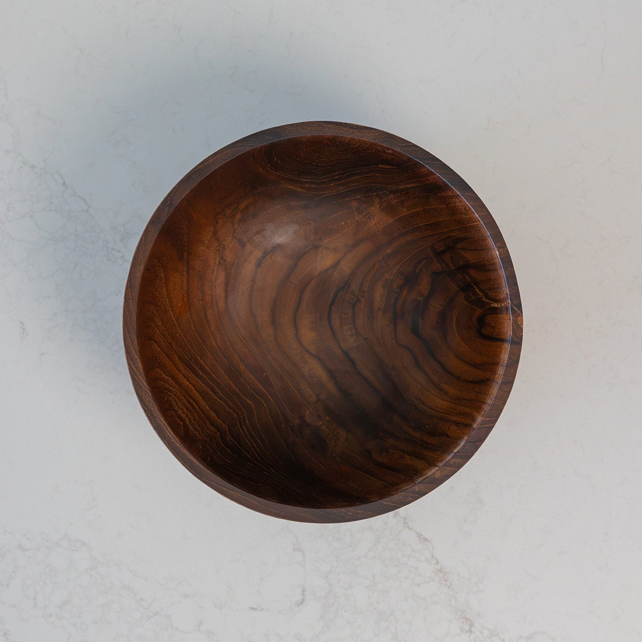 A top-down view of a round wooden bowl with a rich dark brown grain pattern, set against a light gray marble background.