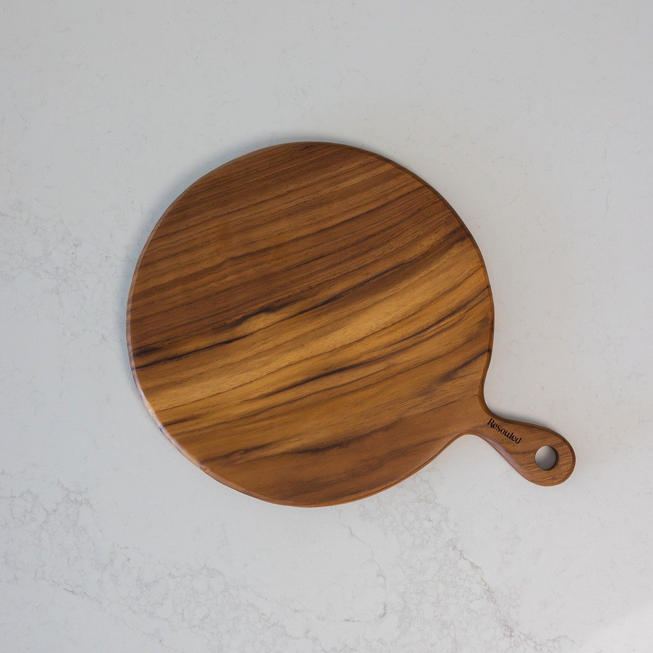A round teak wooden pizza serving board with a handle, featuring a warm medium-brown wood grain pattern, photographed from a top-down angle on a light grey marble surface.