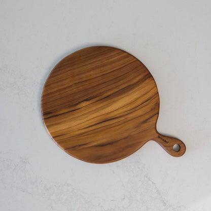 A round teak wooden pizza serving board with a handle, featuring a warm medium-brown wood grain pattern, photographed from a top-down angle on a light grey marble surface.