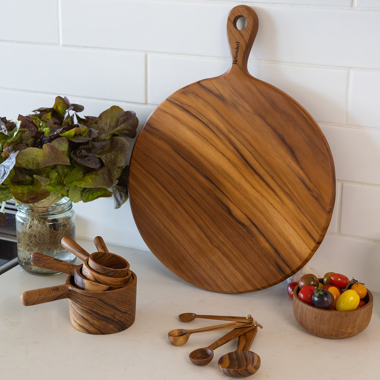 A round teak wooden pizza serving board with a handle, shown on a kitchen counter next to matching wooden measuring cups, spoons, and a small bowl of colourful tomatoes. A white subway tile backsplash is visible in the background.