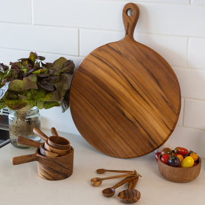 A round teak wooden pizza serving board with a handle, shown on a kitchen counter next to matching wooden measuring cups, spoons, and a small bowl of colourful tomatoes. A white subway tile backsplash is visible in the background.