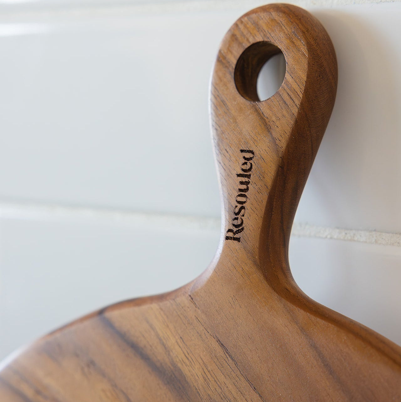 A close-up of a round teak wooden serving board or cutting board with a rounded handle and a circular hole, featuring the engraved brand name 'Resouled' on a white tiled background.