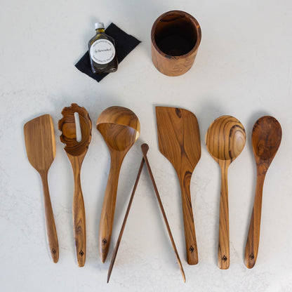 A 8 piece set of sustainable, handcrafted teak kitchen utensils including wooden spoon, spatula, spaghetti spoon, ladle and tongs, displayed next to a small bottle of food safe hemp oil on a light cream background.