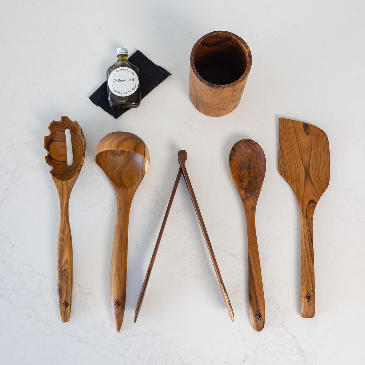A set of six wooden kitchen utensils and a wooden cup arranged on a light gray surface, including a pasta server, soup spoon, mixing spoon, tongs, spatula, and a cup, with a small bottle of balsamic vinegar nearby.