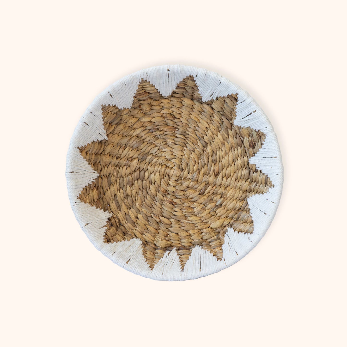 A round woven Water Hyacinth Fruit and Veggie basket with a starburst pattern in natural tan and brown, featuring a white painted rim with small black accents, set against a solid cream background.