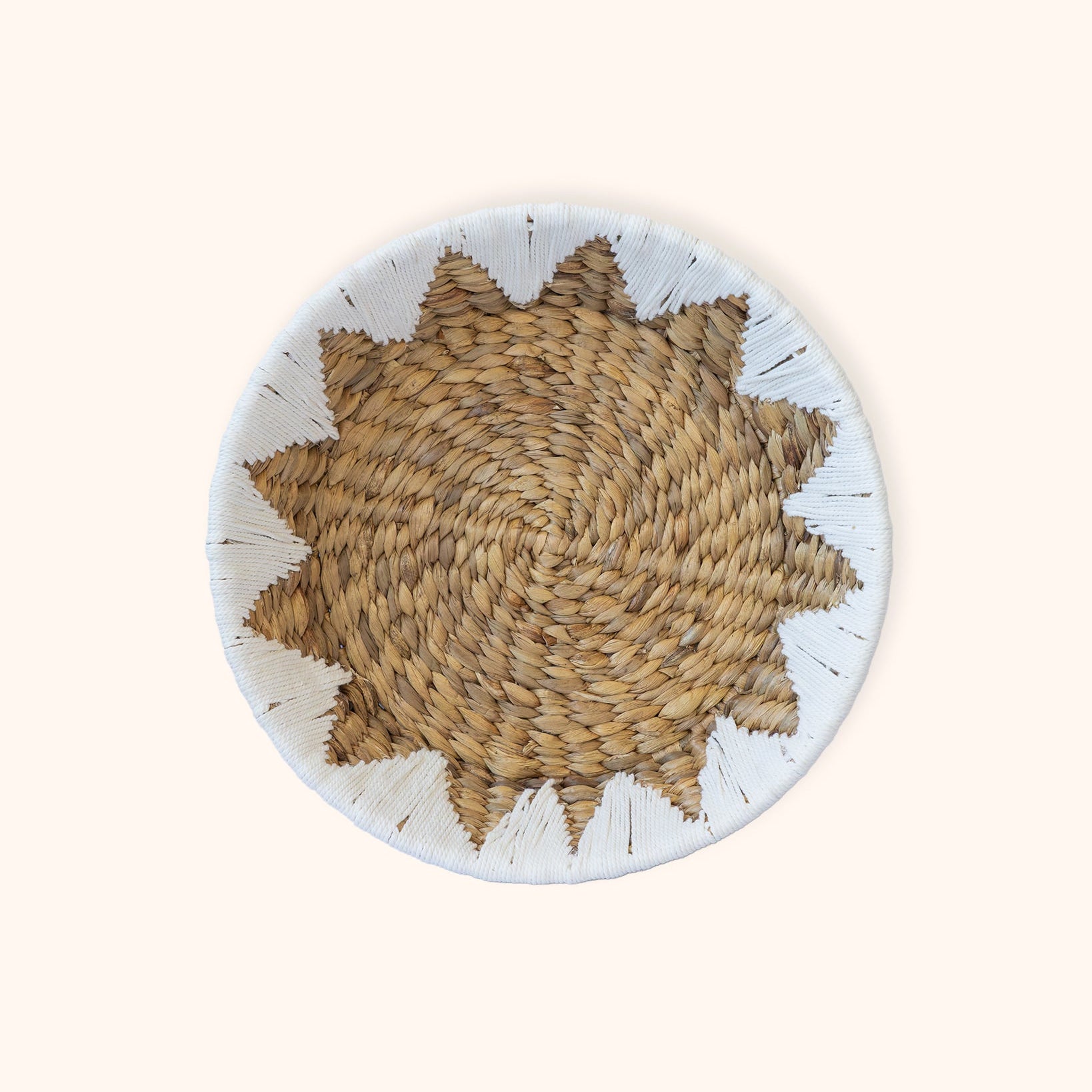 A round woven Water Hyacinth Fruit and Veggie basket with a starburst pattern in natural tan and brown, featuring a white painted rim with small black accents, set against a solid cream background.