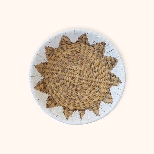 A round woven Water Hyacinth Fruit and Veggie basket with a starburst pattern in natural tan and brown, featuring a white painted rim with small black accents, set against a solid cream background.