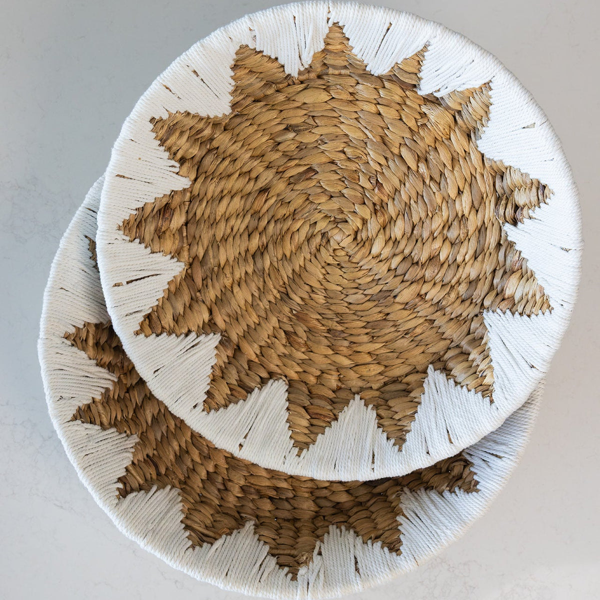 Two round Water_Hyacinth fruit and veggie baskets with a starburst pattern in natural tan and white, stacked on a light gray surface.