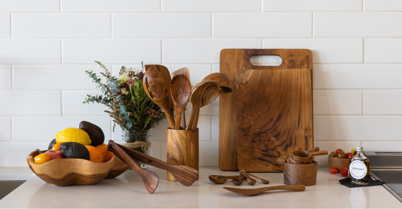 A collection of natural wood kitchen utensils and accessories, including a large cutting board, wooden spoons in a holder, a bowl of fruit, and small wooden containers, arranged on a light countertop against a white subway tile backsplash.