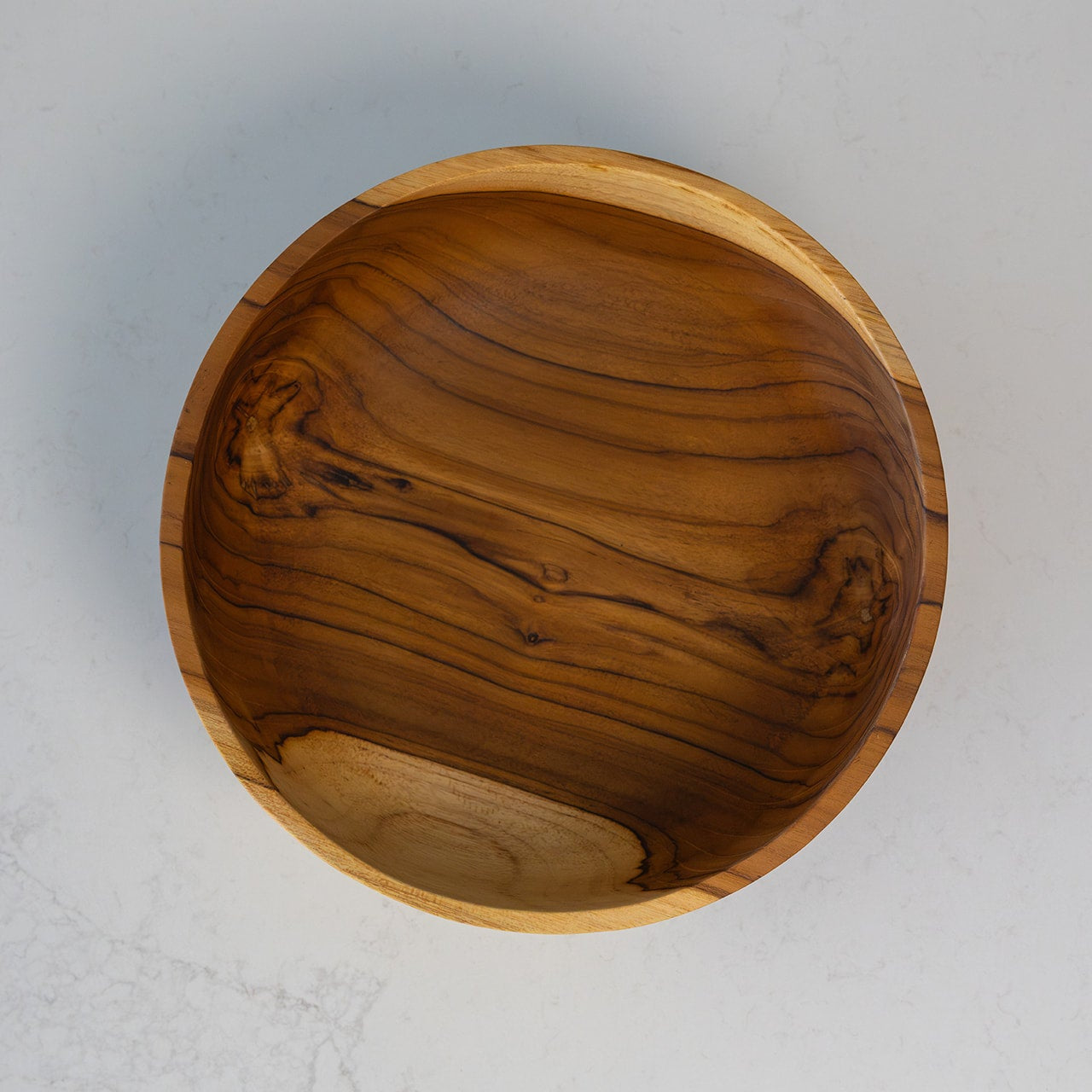 A top-down view of a handcrafted teak wooden bowl with a rich, polished grain pattern, set against a light gray textured background.