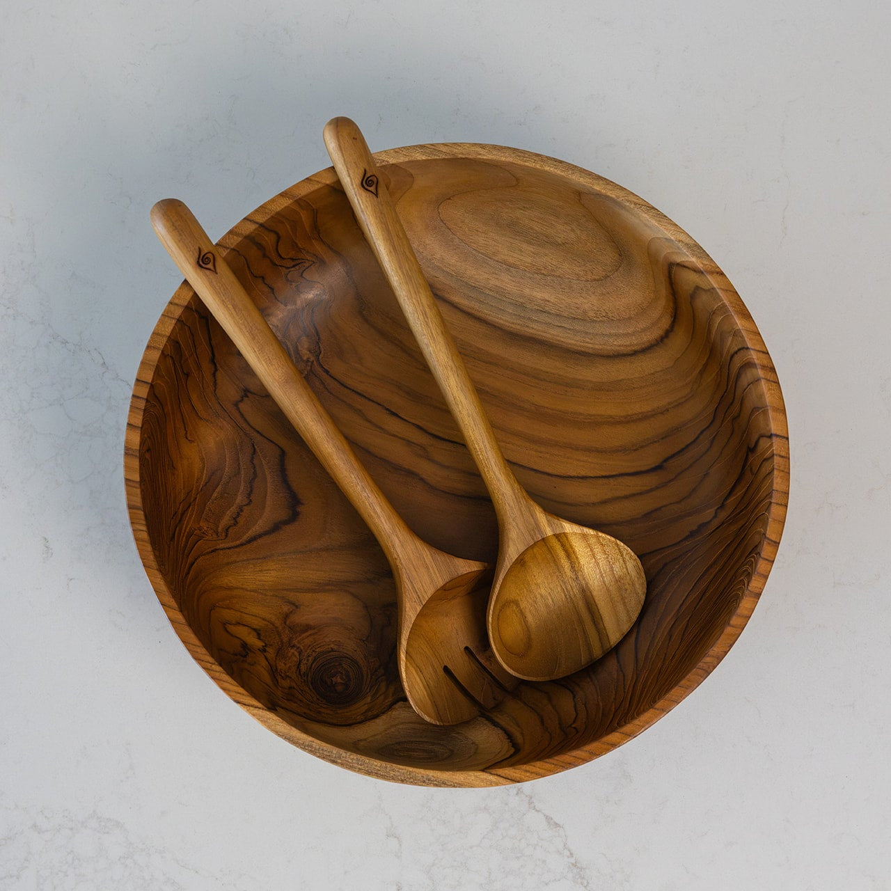 Single piece teak salad bowl with two matching teak wooden salad serving spoons, photographed from above on a marble bench