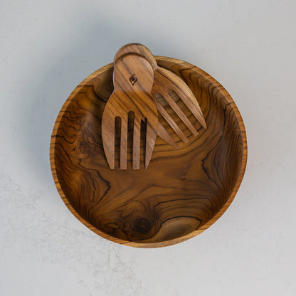 A top-down view of a round wooden salad bowl with a rich dark grain pattern, containing two matching wooden salad server hands on a light gray marble surface.