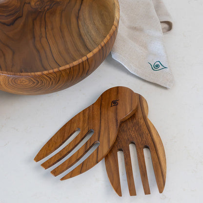 A large teak wooden bowl and matching teak wooden salad hands, arranged on a light beige surface with a folded cream cloth featuring a green logo.