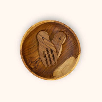 A top-down view of a large round wooden salad bowl containing a pair of wooden salad servers in the shape of hands, all made from teak wood with natural grain patterns, set against a solid off-white background.