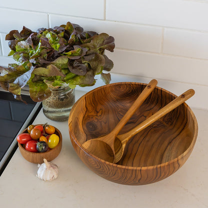 Single piece teak salad bowl with two wooden serving spoons sits on a light countertop. A smaller wooden bowl with colorful cherry tomatoes and a garlic bulb are beside it, with a glass vase of leafy greens against a white subway tile backsplash.