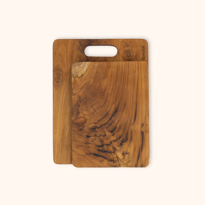 A set of two rectangular teak chopping boards with natural wood grain patterns, one with a cutout handle, displayed on a solid off-white background.