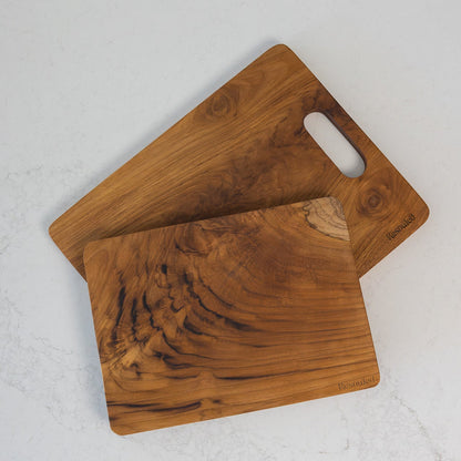 A set of two rectangular teak chopping boards with natural wood grain patterns, one with a cutout handle, displayed on a marble kitchen bench.