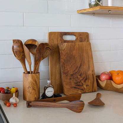 A set of handcrafted teak wood kitchen utensils, including teak chopping boards, rice spoon, and Utensil set, arranged on a light countertop against a white subway tile wall.