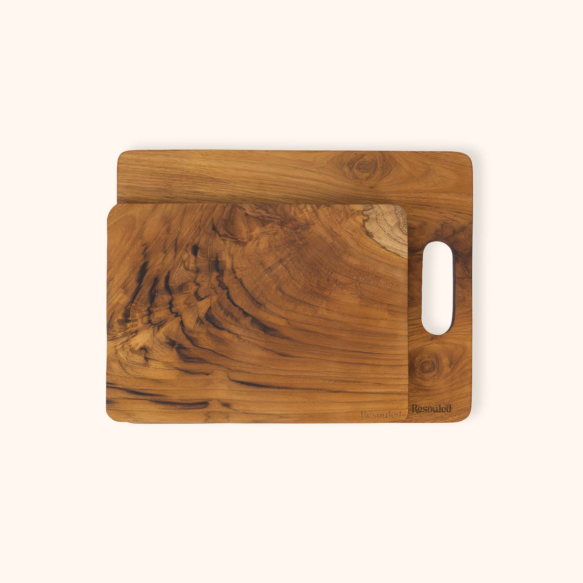 Set of 2 teak chopping boards with natural grain patterns, stacked with the top board offset to the right and featuring a cutout handle on the side, set against a solid off-white background.
