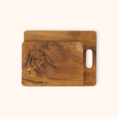 Set of 2 teak chopping boards with natural grain patterns, stacked with the top board offset to the right and featuring a cutout handle on the side, set against a solid off-white background.