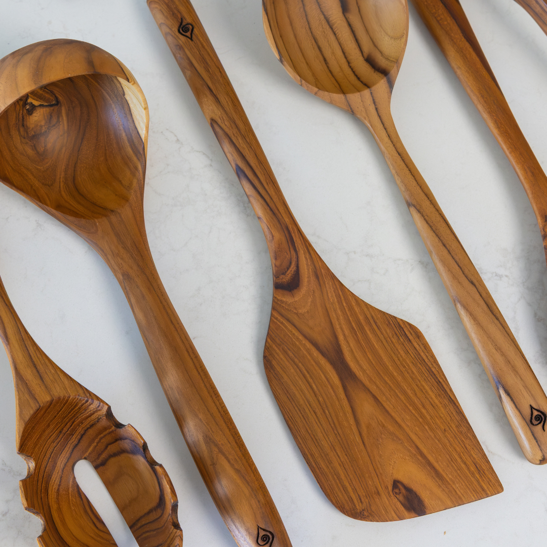 A set of handcrafted teak wood kitchen utensils, including a ladle, spatula, and spoons, arranged diagonally on a light marble surface.