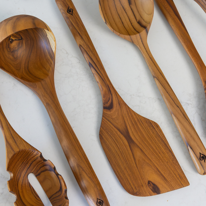A set of handcrafted teak wood kitchen utensils, including a ladle, spatula, and spoons, arranged diagonally on a light marble surface.