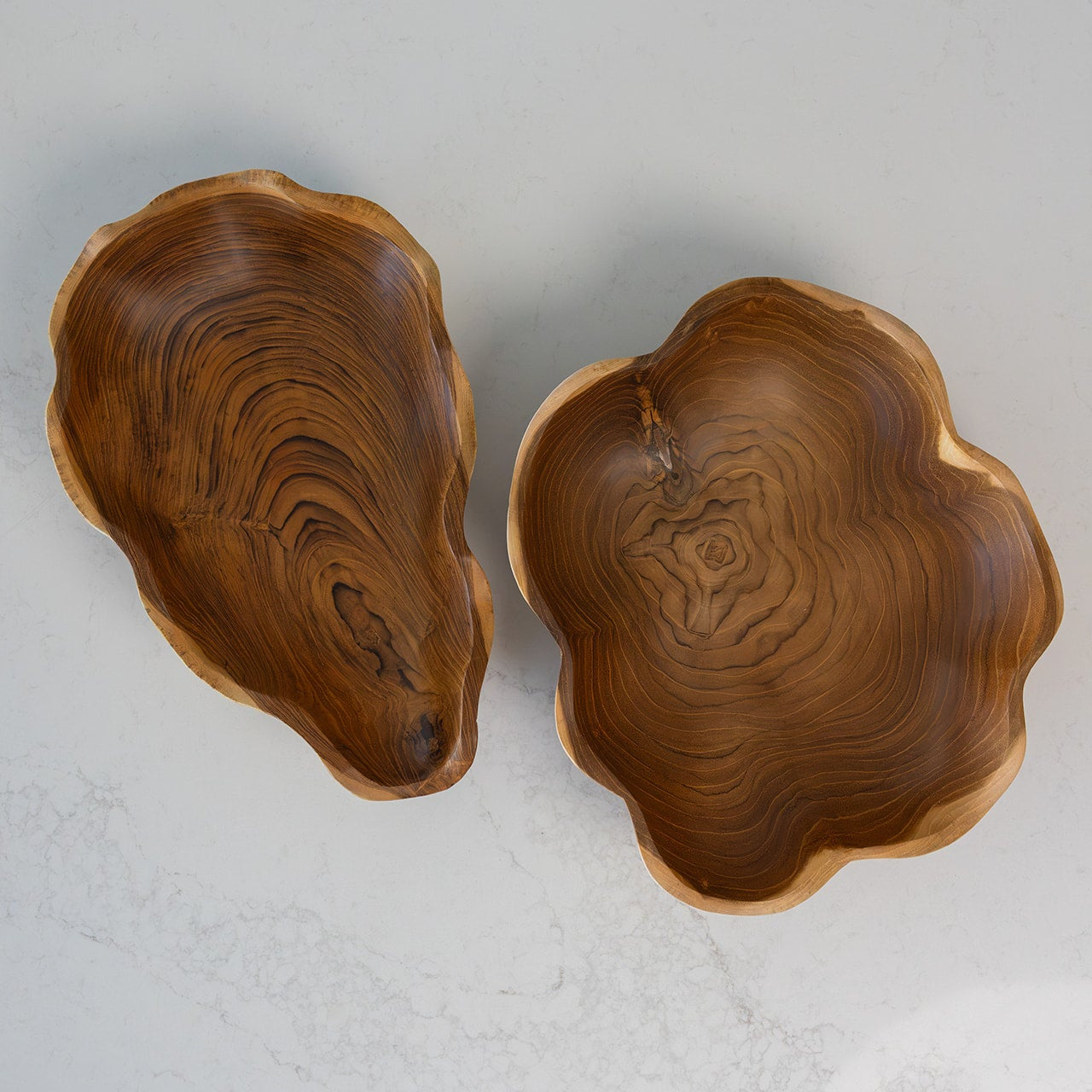 Two organic-shaped teak wooden bowls with natural bark edges, displayed on a light gray marble surface. One bowl is shown from a top-down angle displaying its rich wood grain interior, and the other is shown from a side angle highlighting its depth and irregular rim.
