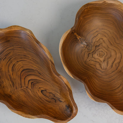 Two organic-shaped teak wooden bowls with rich wood grain patterns, photographed from above on a light gray stone surface.