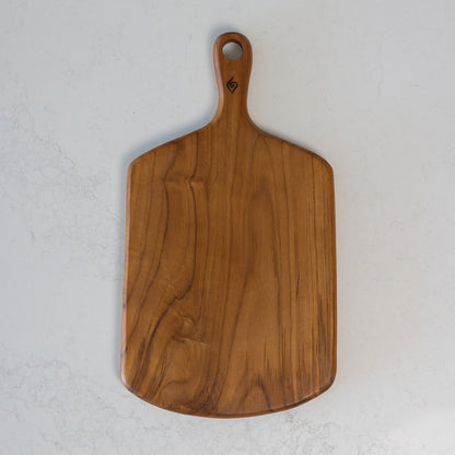 A rectangular teak wooden serving board with rounded corners and a loop handle, shown from above on a light grey marble surface. The board has a warm medium-brown wood grain and a small dark engraved logo near the top of the handle. Ideal for serving cheese and crackers.